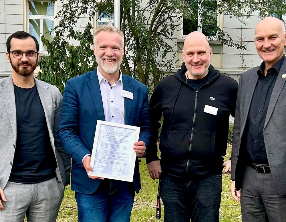 Vier Männer stehen vor einem Gebäude. Einer davom hält ein Zertifikat in den Händen.